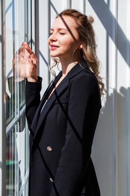 Ein blondes Mädchen in einem schwarzen Anzug steht am Fenster und genießt das Sonnenlicht.