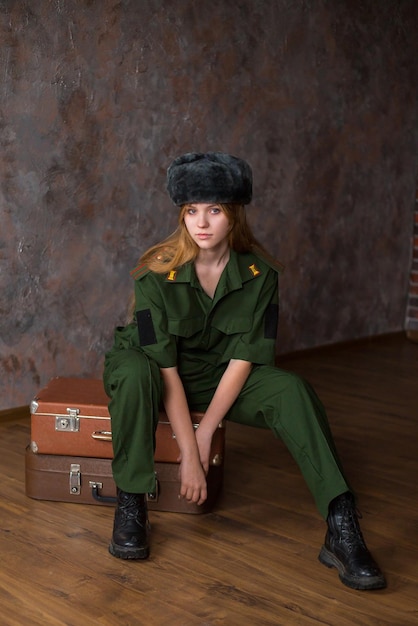 Ein blondes Mädchen in Militäruniform und Wintermütze sitzt auf Koffern vor dem Hintergrund einer Backsteinmauer