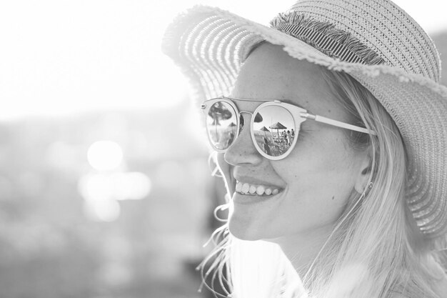 Ein blondes Mädchen lächelnd mit Hut am Strand und Sonnenlicht spiegelt sich in Sonnenbrillen Schöne junge Frauen im Urlaub Porträtfoto