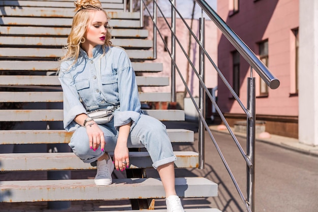 Ein blondes Mädchen mit hellem Make-up und einer Frisur in Denim-Overalls sitzt auf der Treppe auf der Straße