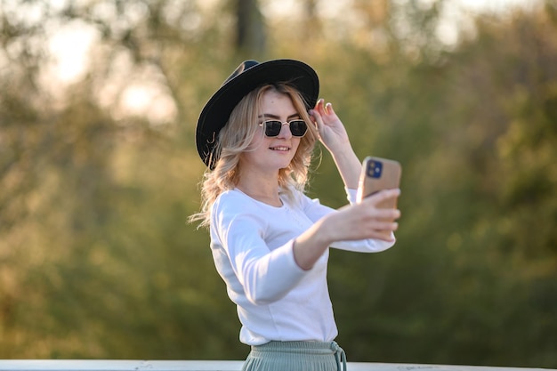 Ein blondes Mädchen mit Hut und Brille schaut im Sommer im Freien ins Telefon