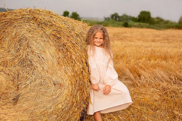 ein blondes Mädchen mit langen Haaren in einem rosa Leinenkleid, das neben einem Heuhaufen auf einem gemähten Feld steht