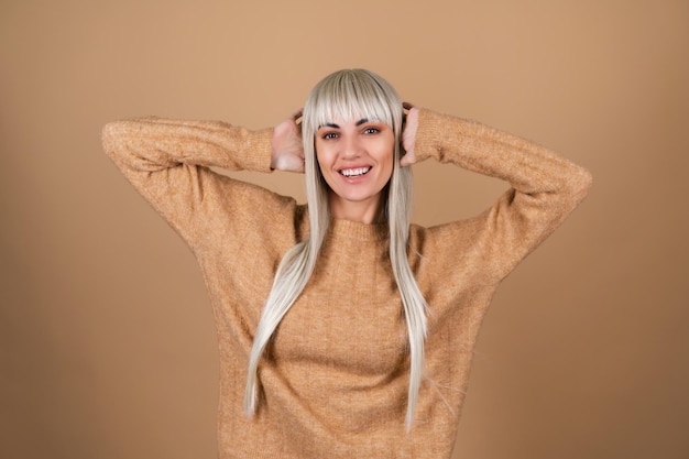 Ein blondes Mädchen mit Pony und braunem Tages-Make-up in einem Pullover auf beigem Hintergrund lacht fröhlich und bedeckt ihre Ohren mit ihren Händen