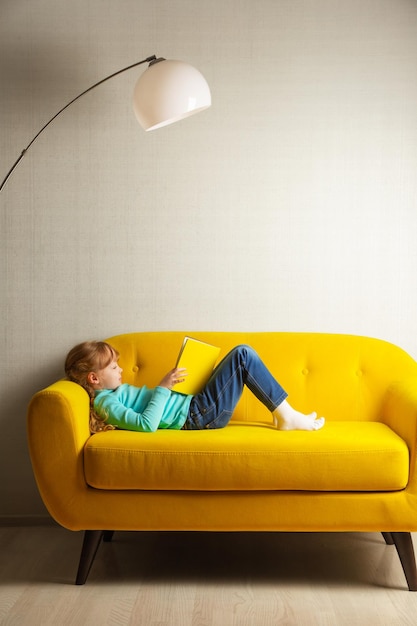 Ein blondes mädchen sitzt auf dem sofa im zimmer und liest ein buch mit gelbem einband