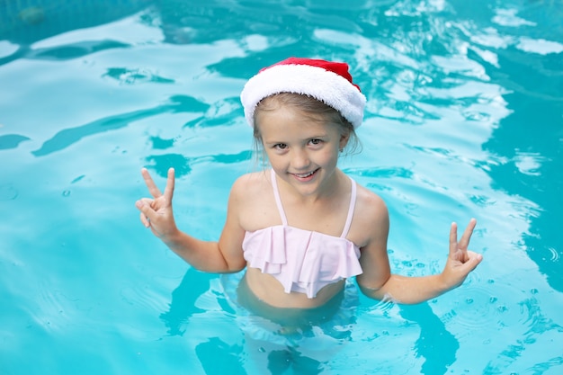 Ein blondes Mädchen steht im Pool in einer rosa Brille mit Weihnachtsmann-Hut und lächelt neues Jahr