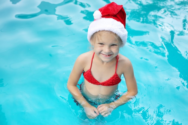 Ein blondes Mädchen steht im Pool in einer rosa Brille mit Weihnachtsmann-Hut und lächelt neues Jahr