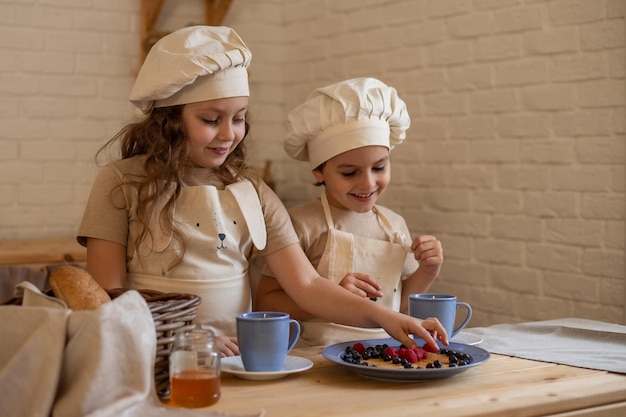 ein blondes mädchen und ein junge in kochmützen und schürzen trinken getränke mit pfannkuchen und beerenlächeln