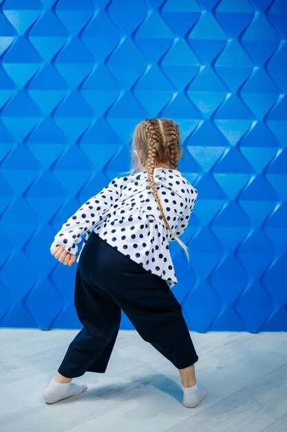 Ein blondes Mädchen von 7 Jahren in einer Jacke mit Erbsen auf blauem Hintergrund tanzt und lacht, helle Kindergefühle der Freude, eine glückliche Kindheit