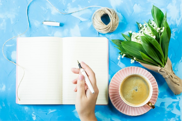 Ein Blumenstrauß aus Maiglöckchen, eine Tasse Kaffee und ein Notizbuch auf einer blauen Tabelle