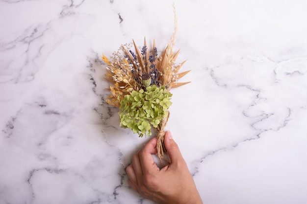 Ein Blumenstrauß aus trockenen Blumen und Kräutern in der Hand einer Frau auf Marmorhintergrund