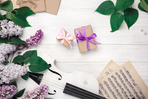 Ein Blumenstrauß von Fliedern mit Violine, Geschenkbox und Musikblatt auf einem weißen Holztisch. Top wiev mit Platz für Ihren Text