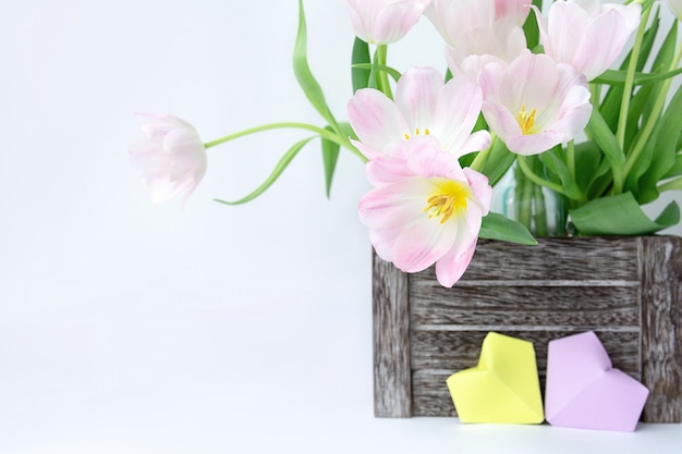 Ein Blumenstrauß von rosa Tulpen in einer Holzkiste und in zwei Papierherzen der gelben und lila Farbe auf einem weißen Hintergrund.