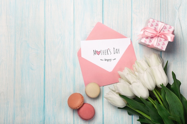 Ein Blumenstrauß von weißen Tulpen mit einer Geschenkbox, macarons, Liebesanmerkung und Farbumschlag auf blauen hölzernen Brettern. Muttertag