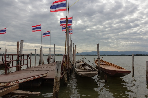 Ein Boot, das an einem hölzernen Pier auf dem Fluss und der Thailand-Flagge bei Kwan Phaya, Thailand gebunden wird