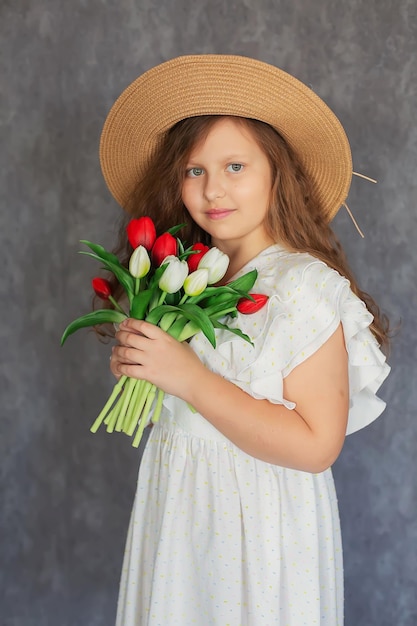 ein braunhaariges Mädchen mit lockigem Haar in einem weißen Kleid und einem Strohhut mit Tulpen in den Händen