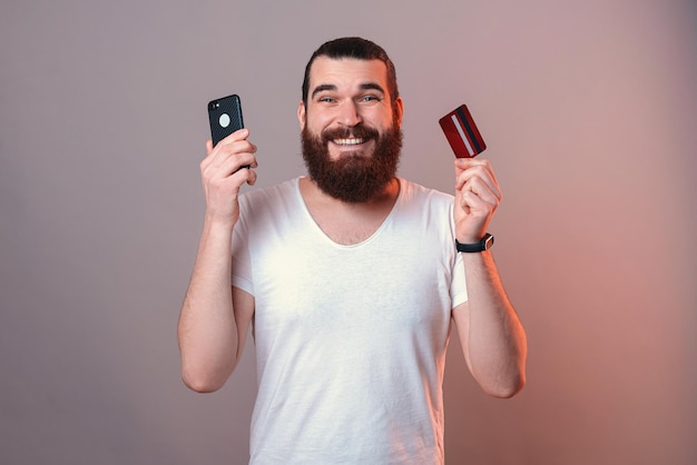 Ein breit lächelnder mann hält eine kreditkarte und ein telefon in der nähe seines gesichts
