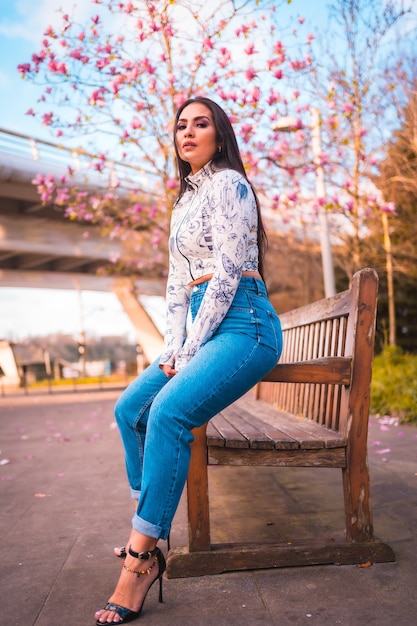 Ein brünettes lateinisches Mädchen, das in einem Park in der Stadt neben einem Baum mit rosa Blumen sitzt