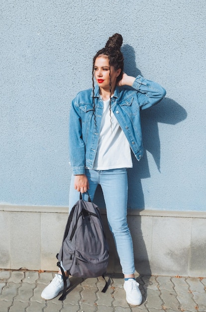 Ein brünettes Mädchen mit Dreadlocks und rotem Lippenstift in einem Jeansanzug steht in der Nähe einer blauen Wand Generation z