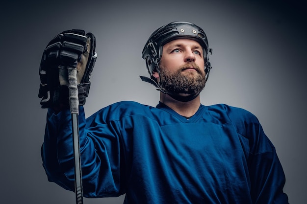 Ein brutaler bärtiger Eishockeyspieler im Schutzhelm hält den Gaming-Stick auf grauem Vignettenhintergrund.