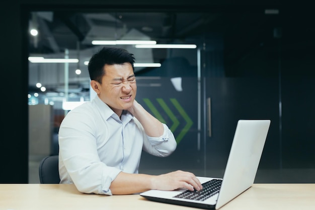 Ein Büroangestellter asiatischer Herkunft arbeitet an einem Laptop und hält sich leidend und windend den Hals