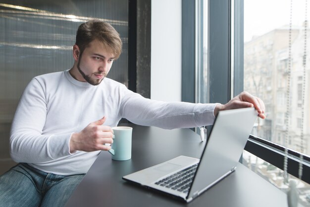 Ein Büroangestellter mit einer Tasse heißem Getränk in den Händen beginnt an einem Laptop zu arbeiten.