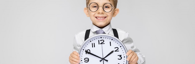 Ein charmanter Junge in einem weißen Hemd, Hosenträgern, einer Krawatte und hellen Jeans steht auf einem grauen Hintergrund. Der Junge hält eine Uhr