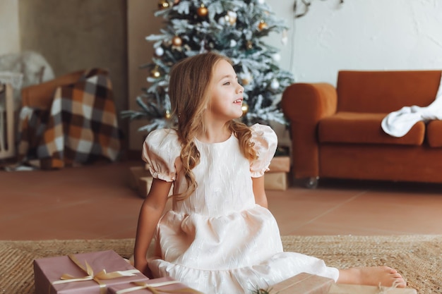 Ein charmantes kleines Mädchen in einem weißen Kleid sitzt zu Hause in der Nähe der Neujahrsdekorationen des Weihnachtsbaums