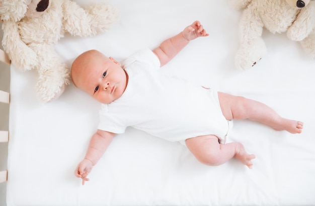 Ein charmantes, lächelndes, blauäugiges, 2 Monate altes Baby in einem weißen Bodysuit liegt in einer Krippe neben einem Teddybär. Blick von oben auf ein Neugeborenes