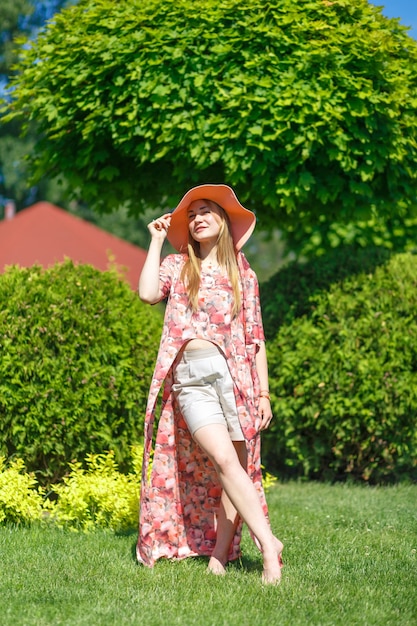 Ein charmantes Mädchen in einem leichten Sommerkleid und einem Pareo-Hut geht in einem grünen Park spazieren.