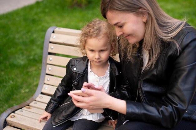 Ein charmantes Mädchen in einer schwarzen Jacke sitzt mit ihrer Mutter auf einer Bank und schaut auf das Telefon