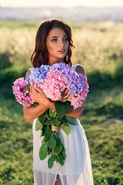 Ein charmantes Mädchen mit einem schönen Lächeln in einem weißen Kleid mit einem Strauß von zarten farbigen Blumen bei Sonnenuntergang.