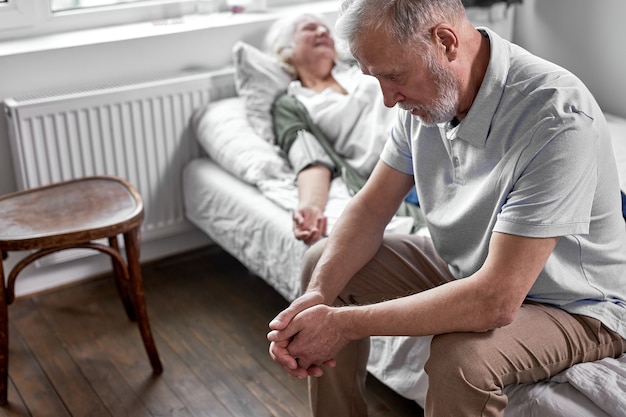 Ein depressiver Mann sitzt in der Nähe seiner kranken älteren Frau und liegt krank im Bett. im Krankenhaus