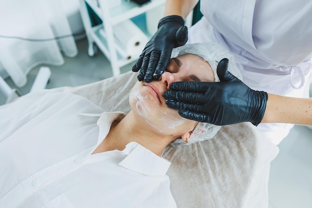 Ein Dermatologe behandelt das Gesicht einer Frau, die auf einem Massagetafel liegt Gesichtshautpflege Eine Schönheitsärztin legt eine Maske auf das Gesicht eines Weibens auf