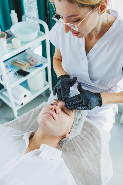 Ein Dermatologe behandelt das Gesicht einer Frau, die auf einer Massageliege liegt. Gesichtspflege. Eine Kosmetikerin trägt eine Maske auf das Gesicht einer Frau auf