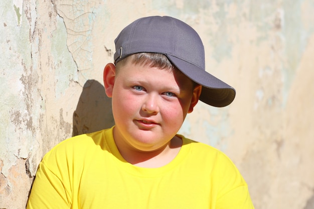 Ein dicker Junge in gelbem T-Shirt und blauer Mütze steht im Sommer an der Wand. Foto in hoher Qualität