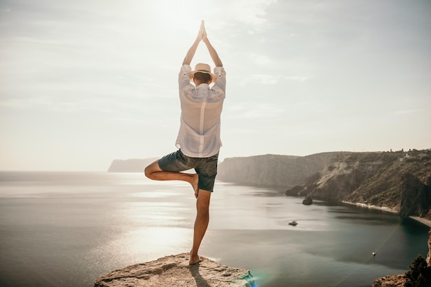 Ein digitaler Nomade, ein Mann mit Hut, ein Geschäftsmann macht Yoga auf den Felsen am Meer bei Sonnenuntergang