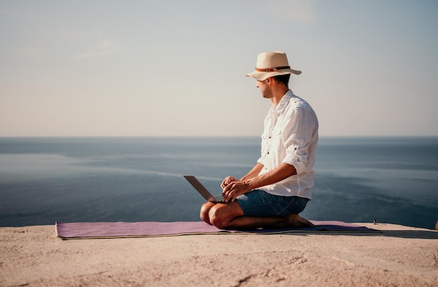 Ein digitaler Nomade, ein Mann mit Hut, ein Geschäftsmann mit einem Laptop macht Yoga auf den Felsen am Meer bei