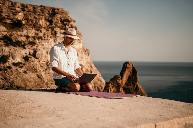 Ein digitaler Nomade, ein Mann mit Hut, ein Geschäftsmann mit einem Laptop macht Yoga auf den Felsen am Meer bei