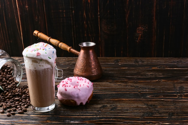 Ein Donut mit rosa Zuckerguss und Schokoladenpulver und ein Cappuccino-Glas mit hohem Schaum und Dekoration. Eine Dose Kaffee und Körner einschenken.