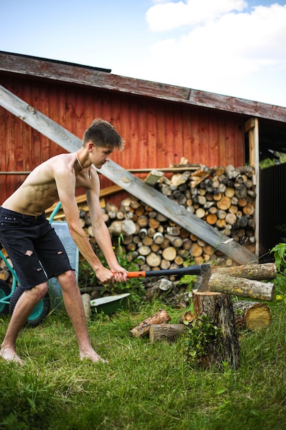 Ein dünner, drahtiger Teenager, barfuß, in schwarzen Shorts, hackt Holz mit einer Axt auf einem Baumstumpf, auf grünem Gras