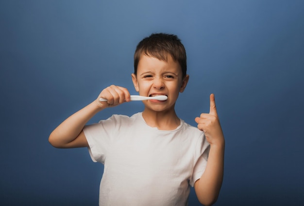 Ein dunkelhaariger Junge in einem weißen T-Shirt auf blauem Grund putzt sich mit einer Zahnbürste die Zähne. Babyhygiene