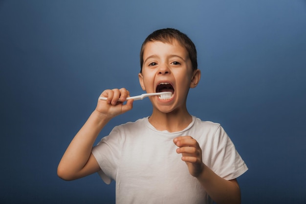 Ein dunkelhaariger Junge in einem weißen T-Shirt auf blauem Grund putzt sich mit einer Zahnbürste die Zähne. Babyhygiene