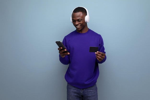 Ein dunkelhäutiger amerikanischer Mann in einem blauen Pullover mit Kopfhörern, einem Smartphone und einer Bankkarte