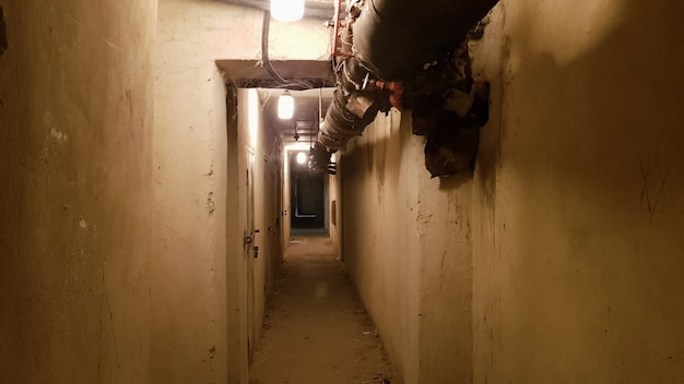 Ein dunkler Keller, ein langer Korridor mit alten rostigen Metalltüren und Rohren. Beleuchteter Tunnel in einem Luftschutzbunker, unterirdischer Militärbunker des Kalten Krieges, Perspektive, getönt.