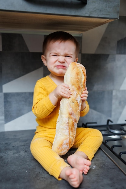 Ein einjähriger kleiner Junge in gelber Kleidung sitzt und isst frisch gebackenes Roggenbrot Das Kind hält ein frisches Baguette in den Händen