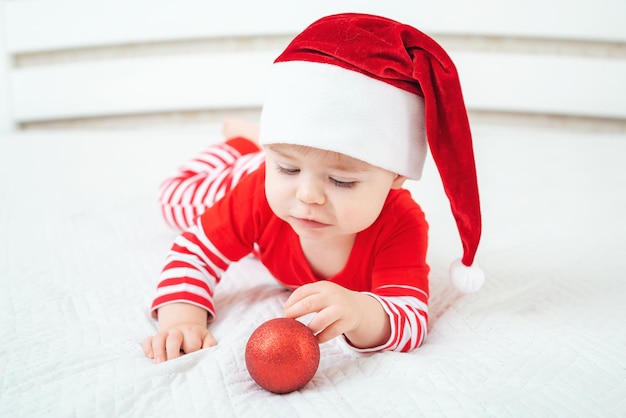 Foto ein einjähriges baby in weihnachtsmütze liegt auf dem bauch und spielt mit einem scharlachroten weihnachtsballkind, das rot trägt