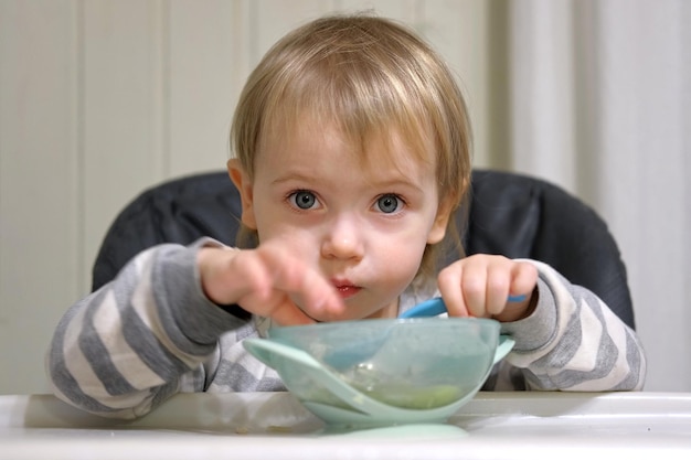 Ein einjähriges hungriges Mädchen in gestreifter Freizeitkleidung sitzt im Hochstuhl am weißen Tisch und isst selbst Reis mit einem Löffel. Unscharfer Hintergrund im Esszimmer. Gesunde Ernährung für Kinder. Kinderernährung