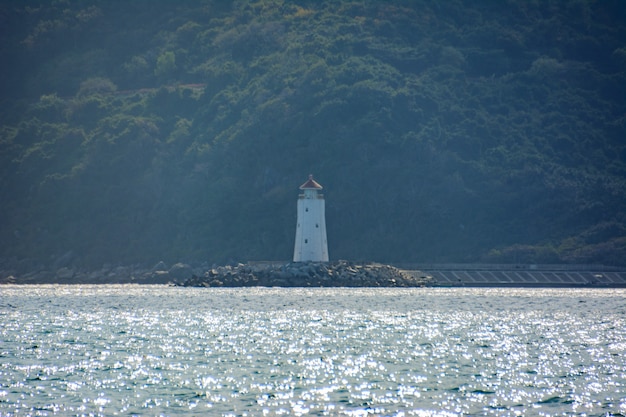 Ein einsamer Leuchtturm ist im Nebel vor dem Hintergrund von Regenwald, Bergen und klarem türkisfarbenem Meer an der Küste der Yalong-Bucht im Südchinesischen Meer zu sehen. Sanya, Insel Hainan, China. Natur Landschaft.