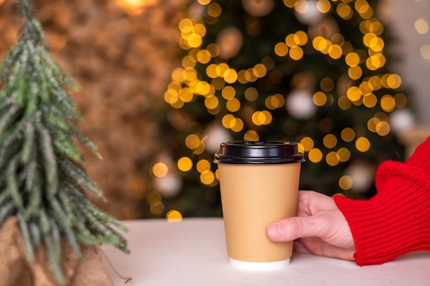 Ein Einwegbecher, um eine Hand in einen roten Pullover auf dem Tisch neben dem Weihnachtsbaum zu legen