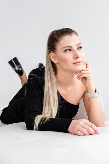 Ein elegantes Mädchen in einer schwarzen Bluse mit einer offenen Schulter, einer intelligenten Uhr auf dem Arm, einem hohen Pferdeschwanz und Make-up liegt auf dem Sofa und posiert für die Kamera